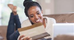 young woman with a smile on her face laying on her couch in front of her laptop holding a small brochure in her hand